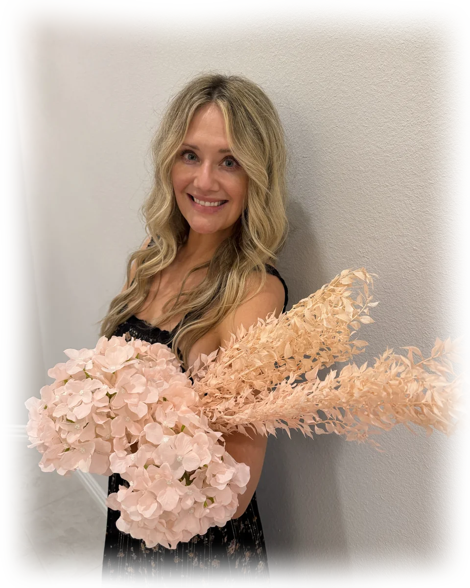Nikki Nielsen, founder and lead floral designer of the Petite Petal team, smiling and holding blush hydrangeas and soft peach foliage